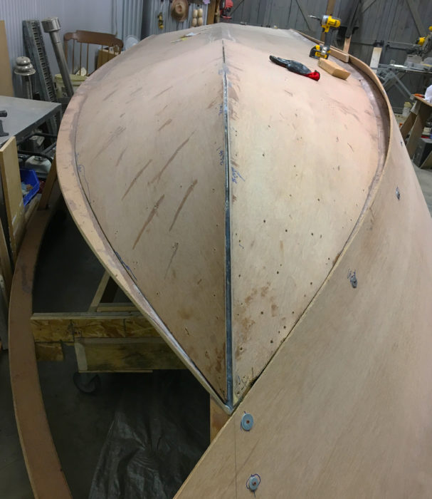At this stage of the construction, the bottom panels have been assembled, including the second layers of 1/4” plywood at the bow and the chine flats. The 4” chine flat at the left is clearly visible at the edge of the bottom panel. The first side panel has been installed with the help of screws and fender washers.