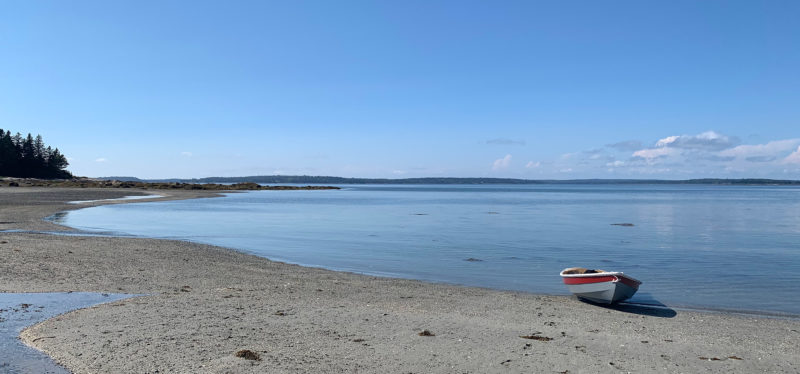 The unrigged shellback dinghy lays on a beach at mid-tide. The water has started to go out and has exposed the gray sandy beach of Pond Island.