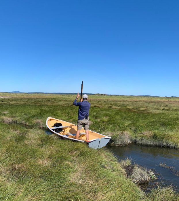 Tom stands in the shellback and uses an oar to punt the boat through a narrow stream