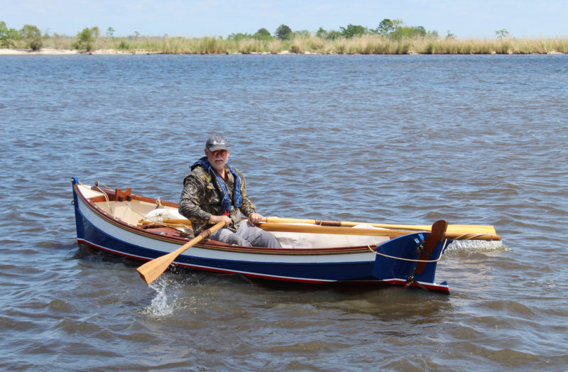 A man (in a lifejacket obviously) rows the Little Crab from the center thwart. The skiff is balanced on the calm water.
