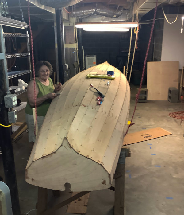 The 15-foot Chester Yawl is supported by sawhorses and lines from the basement roof. The boat's hull is completed and Dennis's mom smiles while examining it... possibly she, too, is impressed that the boat fit in the basement.