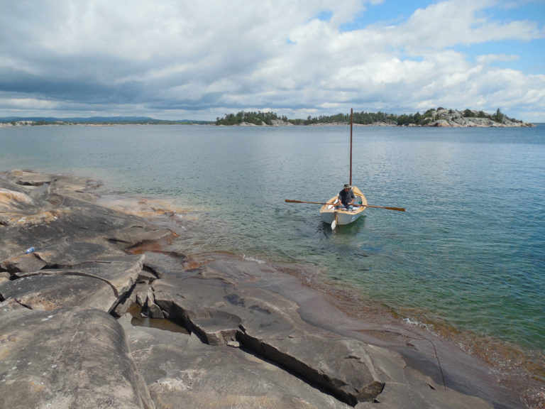 Sailor rows his small boat near a rocky shore with an island in the background.