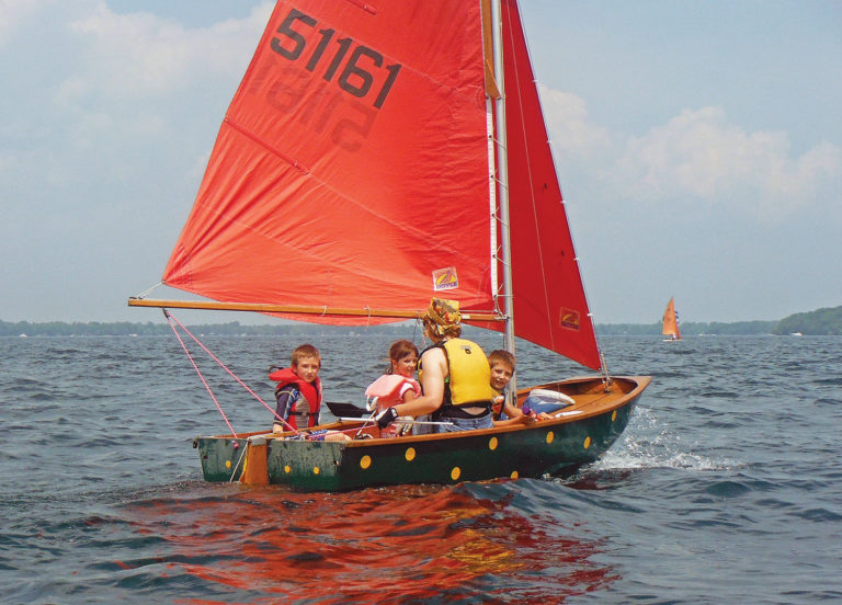 Adult sailor and three kids all wearing live vests in a small sailboat with red sail.