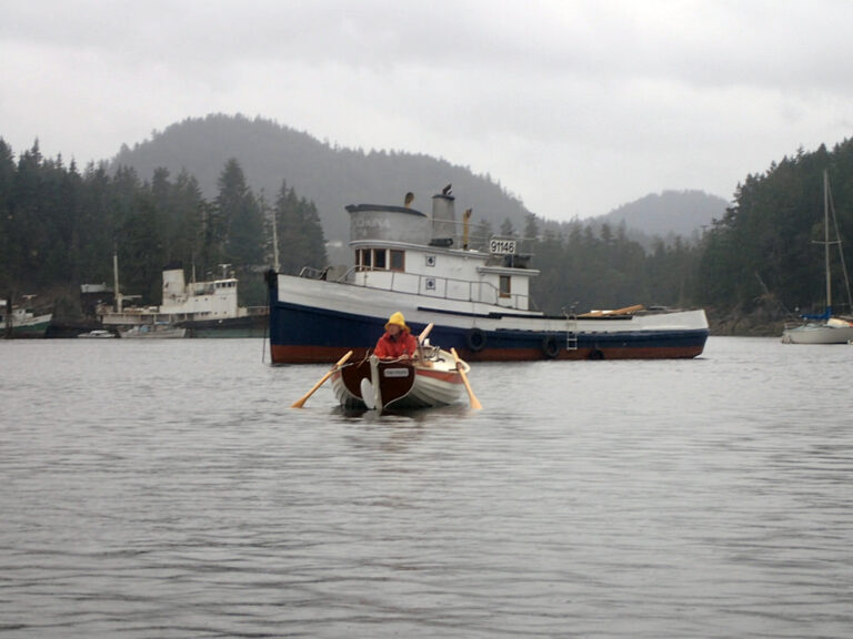 Man in rain gear rows a small wooden boat in a harbor near other larger boats.