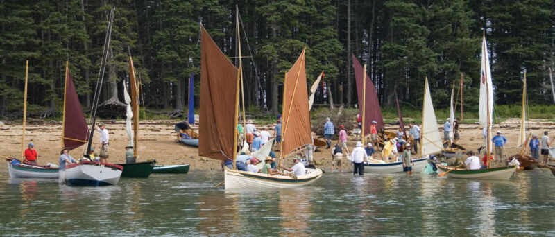 Various small boats sit docked on a beach at the Small Reach Regatta as people help to bring them in or launch them.