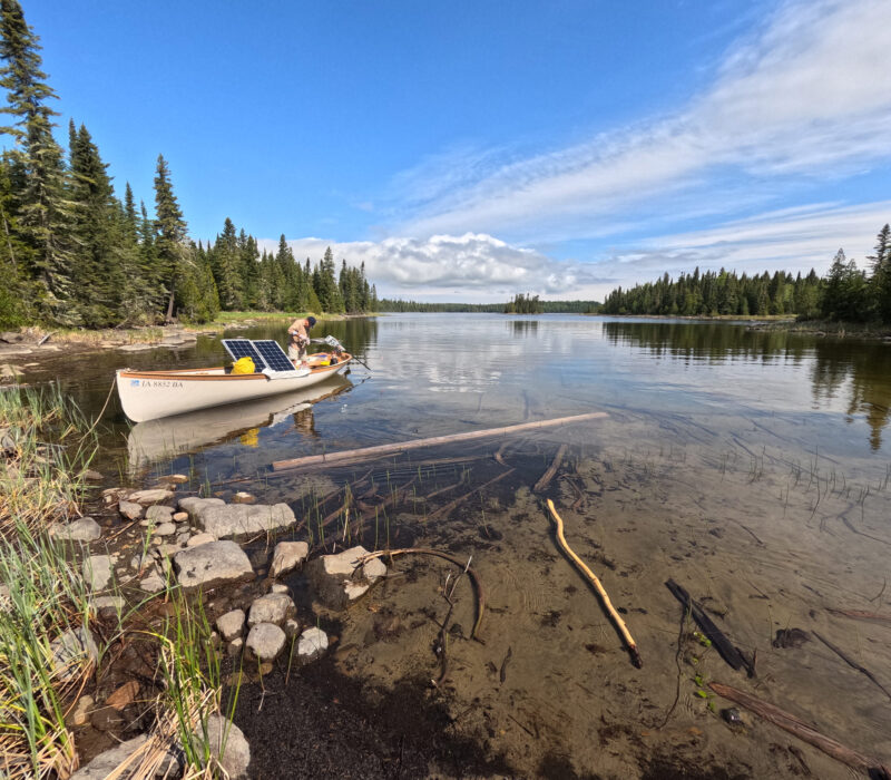 Man stands aboard a white boat with solar panels near the shore of Fishhook Island while cruising Lake Nipigon.