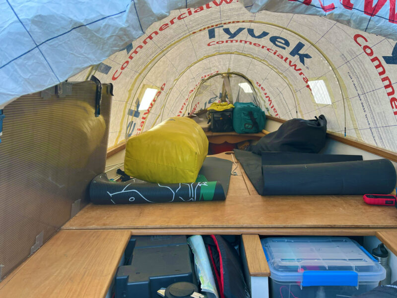 A boat's interior with bunks and stowed gear sheltered by a makeshift Tyvek tent.