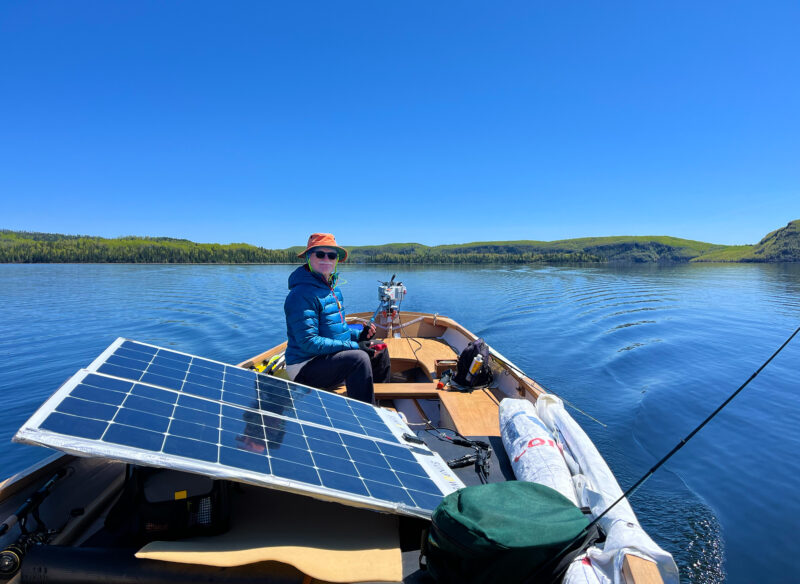 Man sits aboard a boat next to a solar panel under a clear blue sky.