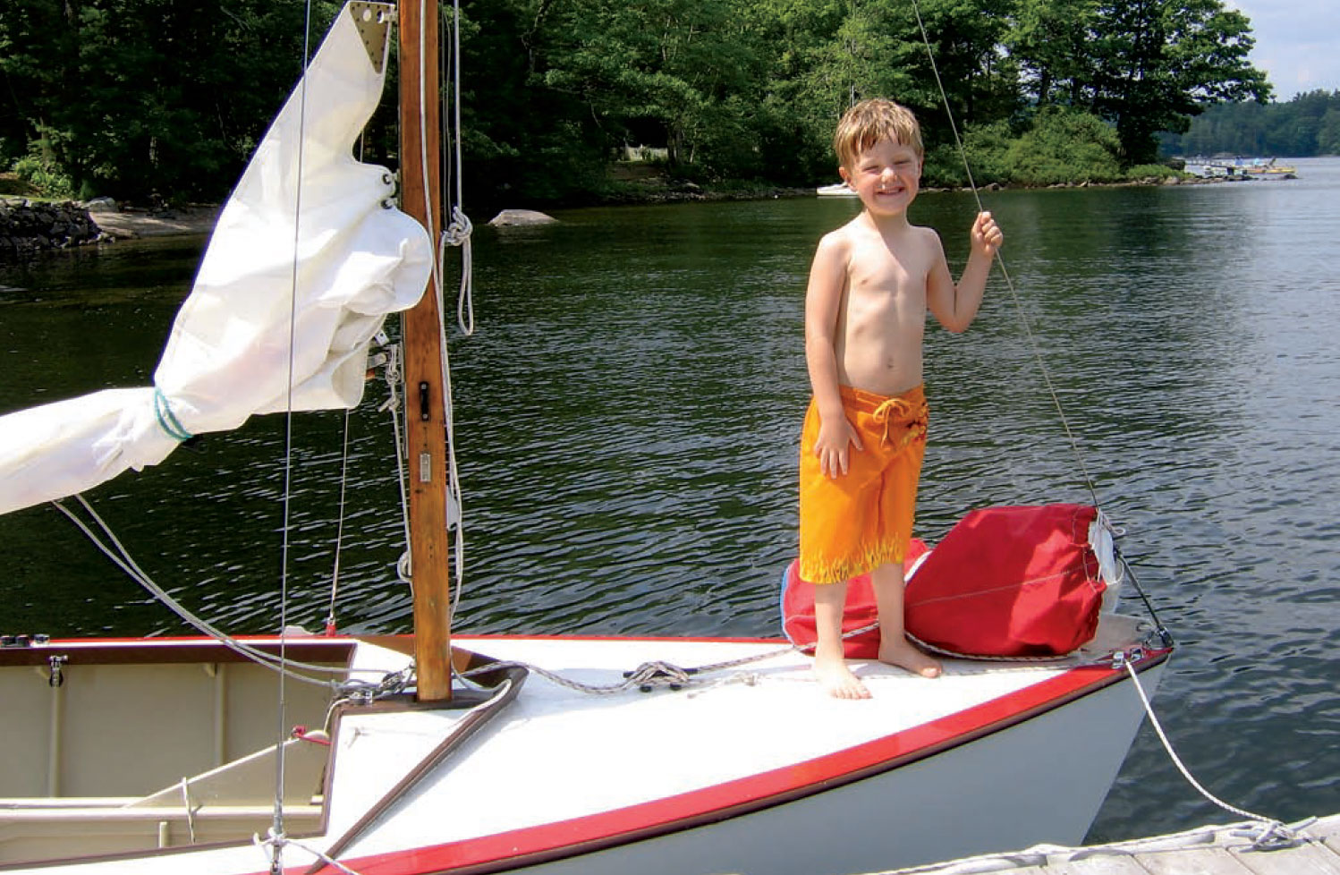 Little boy in swim trunks stands on the bow of a small sailboat.