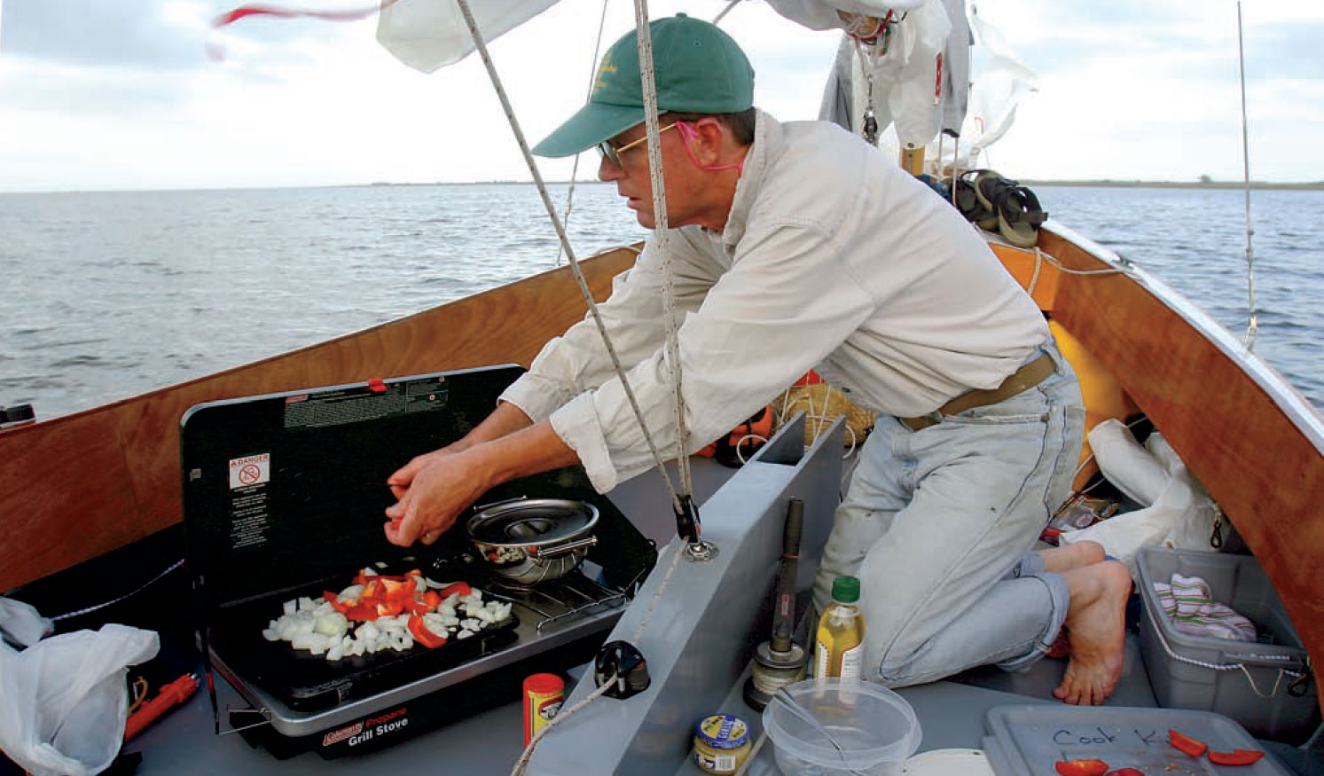 Man cooks food with small portable stove aboard a sailboat.