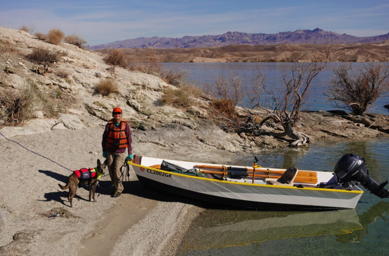 Woman and dog with small open boat beached on cold day.