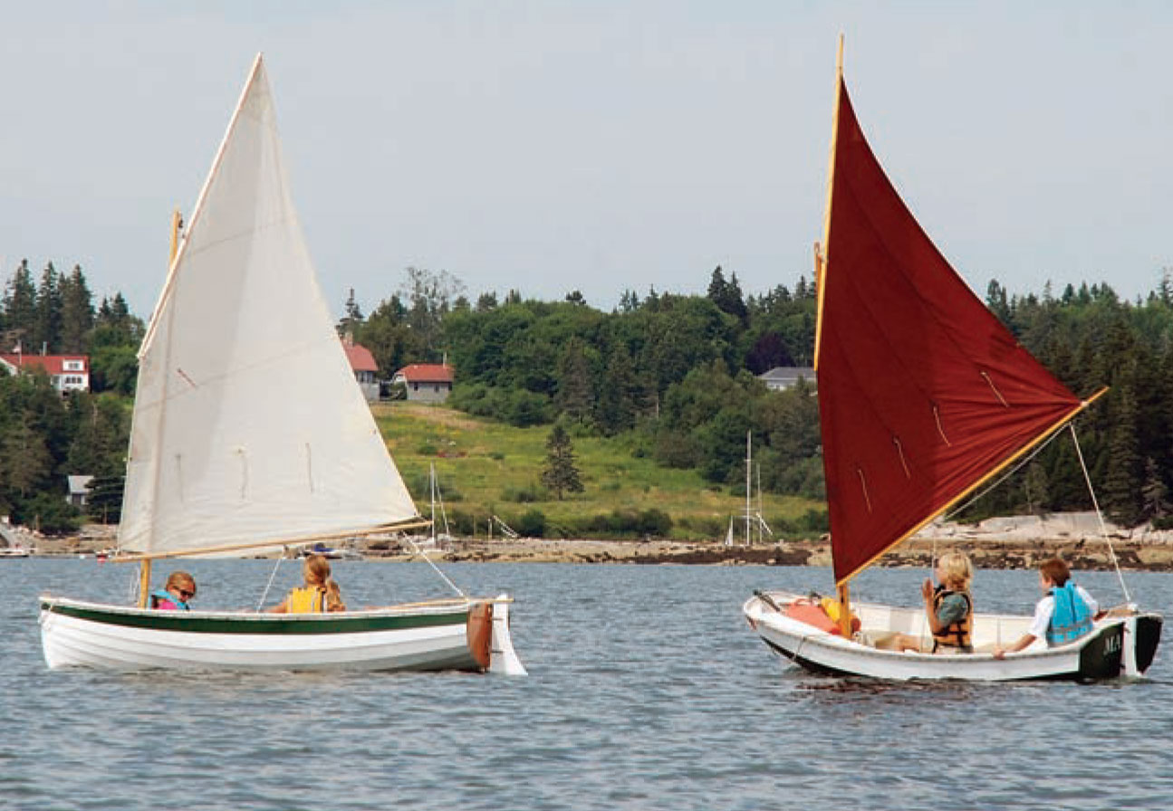 Kids sail a Drifter sailboat and Nutshell pram in the water with a wooded shore in the background.