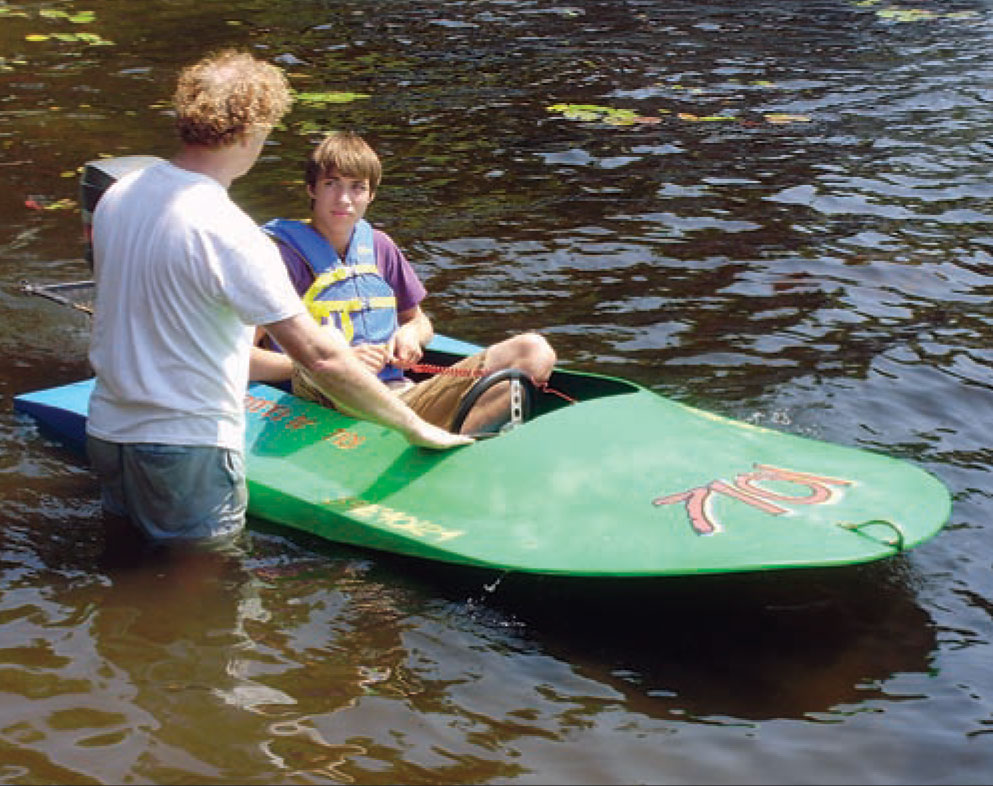 Man stands nearly waist-deep in water while talking to a young man sitting in a blue and green Sea Flea power boat.