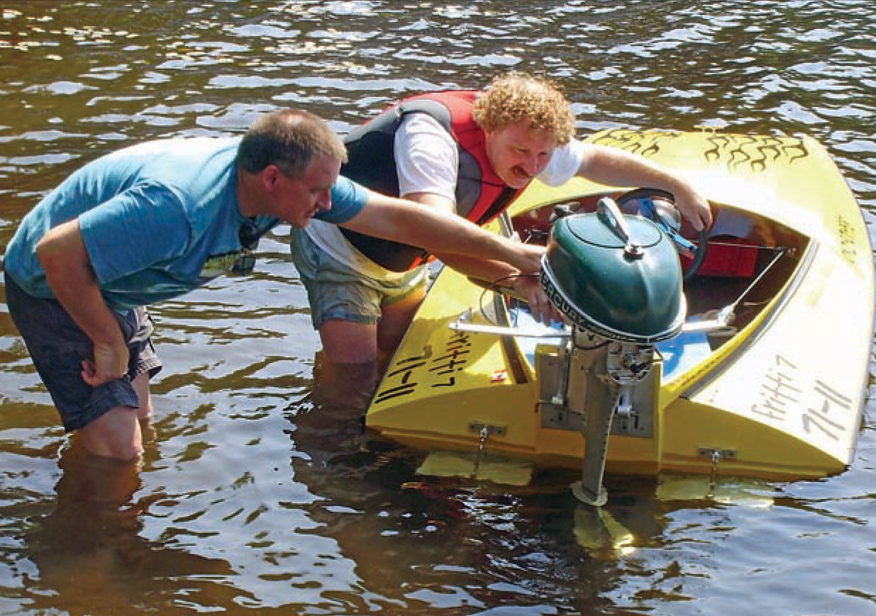 Two men inspect the outboard motor of a yellow Sea Flea powerboat.
