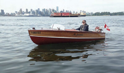 Man wearing a ball cap and sunglasses sits in a small wooden powerboat in a harbor with view of a city and commercial vessels in the background.