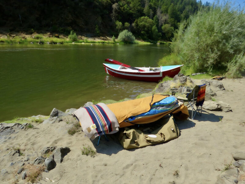 Cot with bedroll beside a river and a moored wooden river boat.