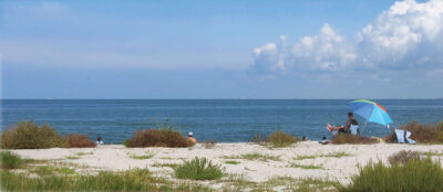 Man sits under a colorful umbrella on a sandy beach.