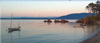 Three small sailboats in the water near a rocky shore with mountains in the background at sunset.
