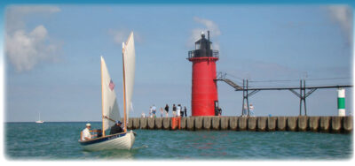 Sailor cruises in a small white sailboat in front of the South Haven South Pierhead Light in Michigan.
