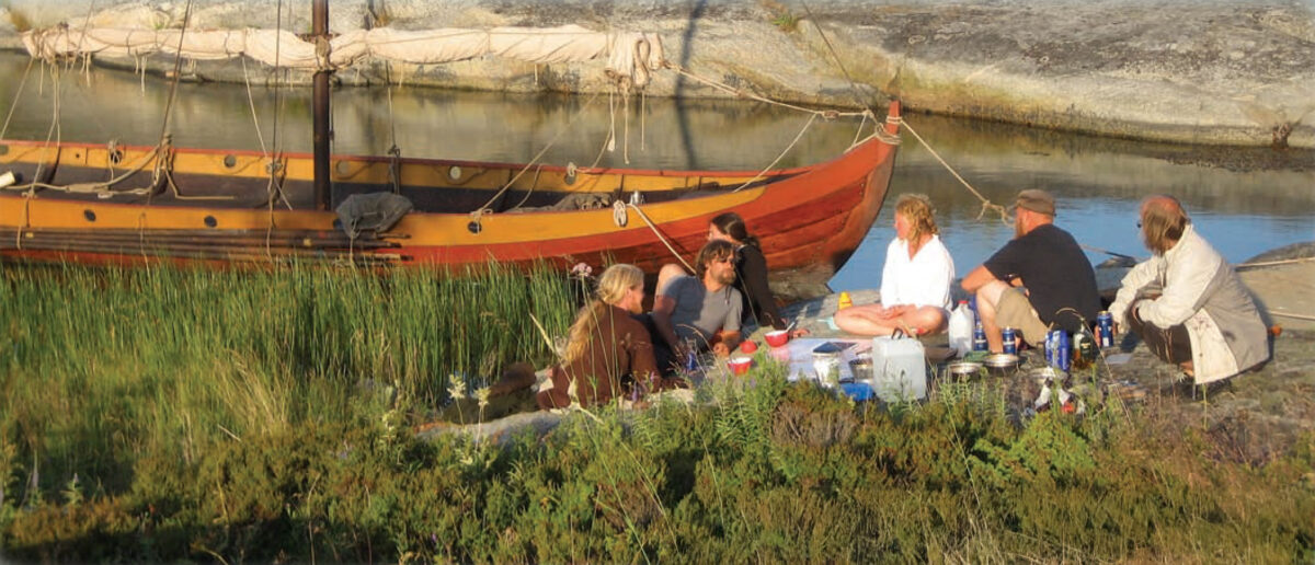 Group of people sit at their campsite next to a wooden sailboat.