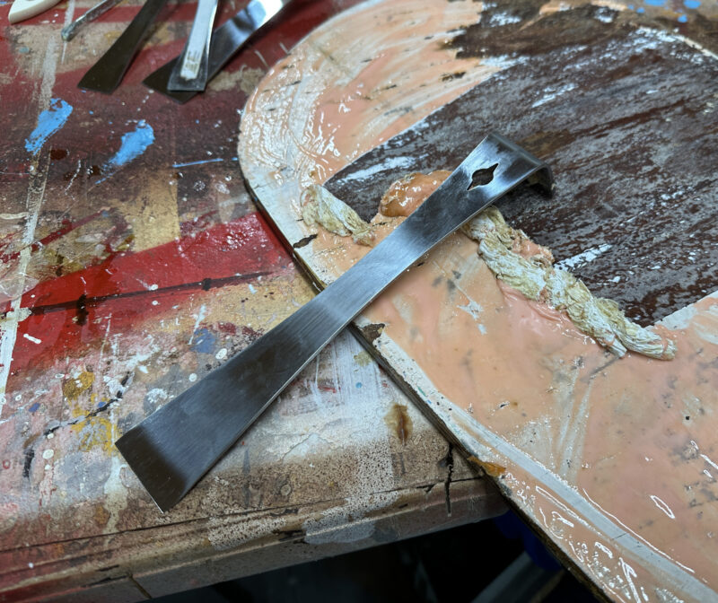 Stainless-steel scraper bar removing finish on a centerboard.