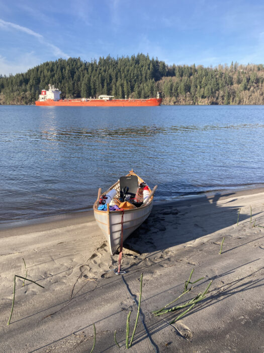 Red-hulled freighter traveling upstream on Columbia River; skin-on-frame boat on beach in foreground.