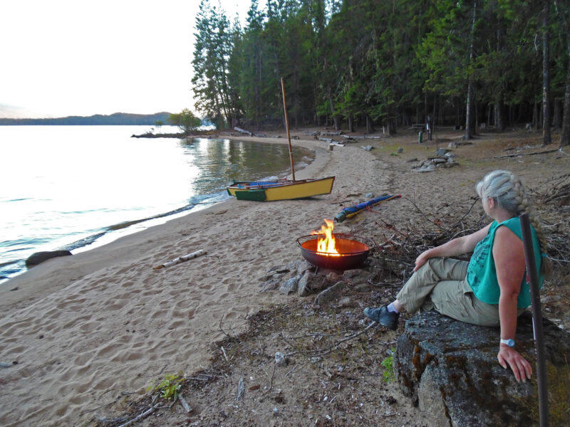 A woman looks at a firepit with a small boat pulled up on sandy beach in the background.