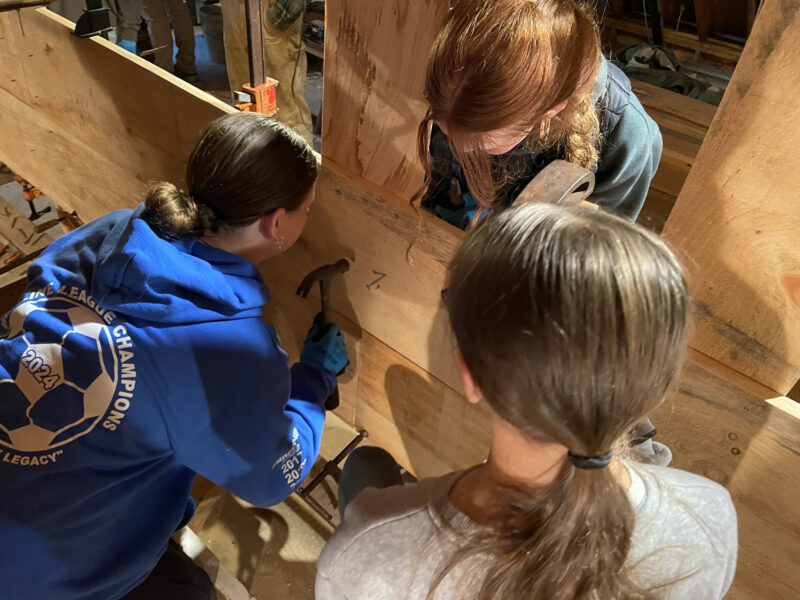 Young students hammer nails into a boat’s bottom board during construction.