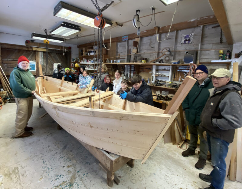 A group of students and boatbuilders gather around a partially built bateau.
