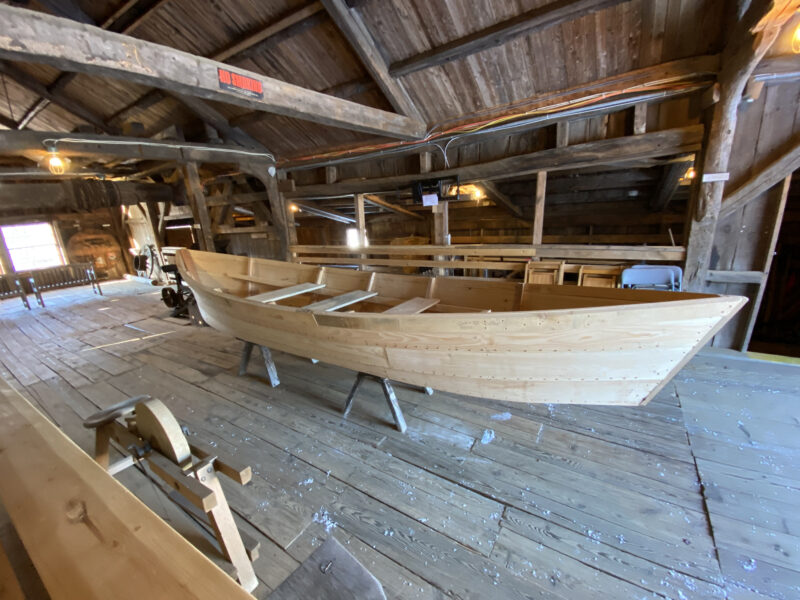 A finished bateau sits on saw horses in a timber-frame workshop.