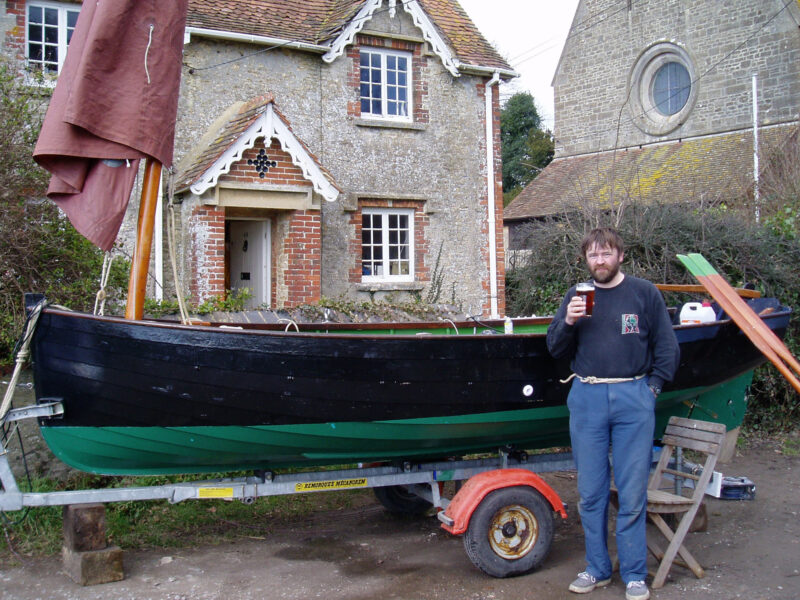 Man standing in front of black-painted Ilur sailing dinghy on a trailer.