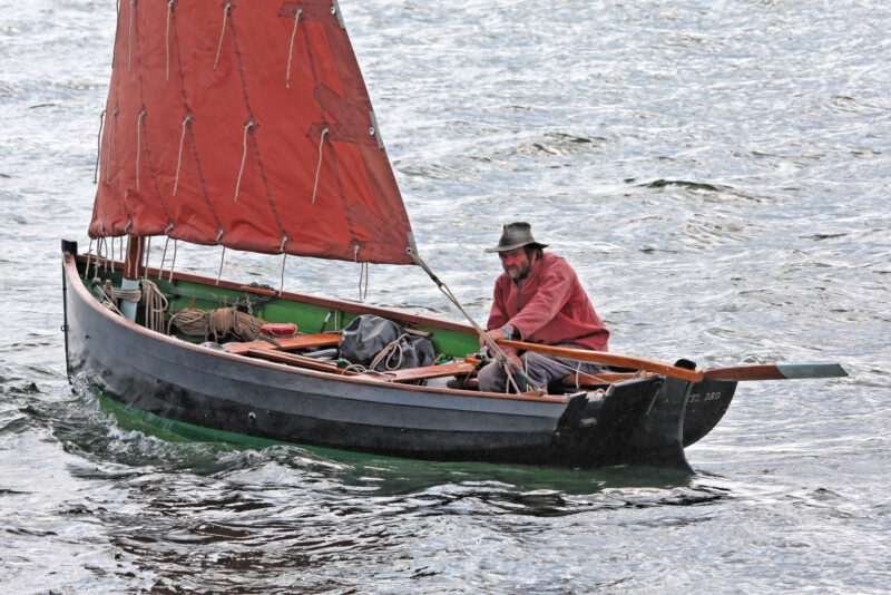 A man in a hat and Breton-red smock looks under his open boat’s tan-bark sail.