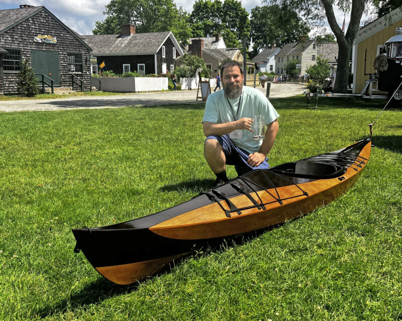 Man kneeling on grass beside a varnished stitch-and-glue kayak.